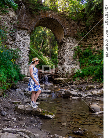 Old stone arched destroyed bridge over small river in a mountain forest 109183407