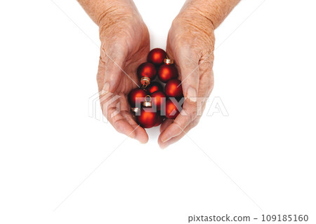 Happy holidays. Old wrinkled female hands hold beautiful red Christmas toys in palms on white background. Elderly people stay at home alone on Christmas holidays concept. Copy space. Top View Happy holidays. Old wrinkled female hands hold beautiful red Christmas toys in palms on white background. Elderly people stay at home alone on Christmas holidays concept. Copy space. Top View 109185160