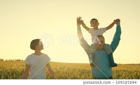 a man walks through a field with wheat with a small son child on his shoulders. cheerful happy family at sunset. happy family life. child loves parents mom dad. walk trip weekend together. smiling a man walks through a field with wheat with a small son child on his shoulders. cheerful happy family at sunset. happy family life. child loves parents mom dad. walk trip weekend together. smiling 109185894