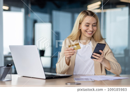 Young joyful woman at workplace inside office holds phone , bank credit debit card in hands, businesswoman chooses gifts in online store, book service and transfer funds using app on smartphone. 109185983
