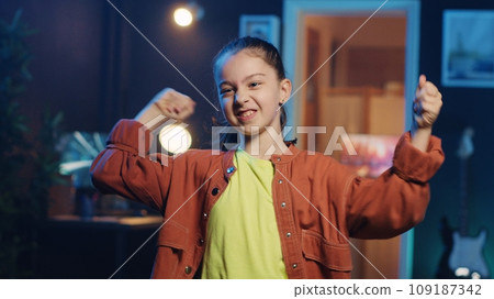 Close up shot of small girl doing captivating dance moves for social media platform, doing viral challenge. Cute little kid recording video for online fans in dimly lit home studio Close up shot of small girl doing captivating dance moves for social media platform, doing viral challenge. Cute little kid recording video for online fans in dimly lit home studio 109187342