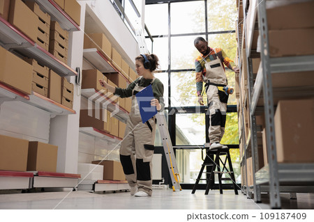 Storehouse worker analyzing cardboard box details, working at goods delivery for customers in storage room. Woman supervisor wearing headphones listening music while preparing merchandise Storehouse worker analyzing cardboard box details, working at goods delivery for customers in storage room. Woman supervisor wearing headphones listening music while preparing merchandise 109187409