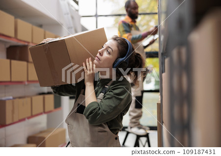 Woman manager holding cardboard box during inventory, working at customers orders preparing merchandise for delivery. Storage room supervisor wearing headphones listening music in warehouse Woman manager holding cardboard box during inventory, working at customers orders preparing merchandise for delivery. Storage room supervisor wearing headphones listening music in warehouse 109187431