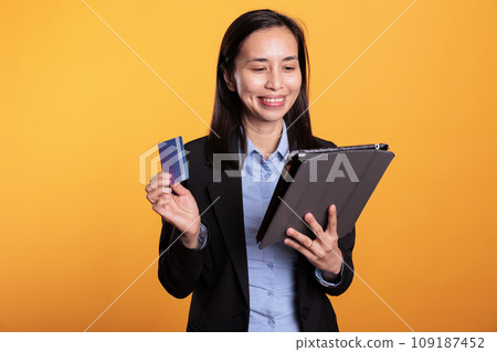 Cheerful model using credit card to do online shopping, ordering stylish clothes using tablet computer. Asian attractive woman making electronic payment transaction on retail store website 109187452