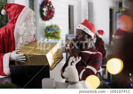African american employee surprised to see Christmas gift offered by coworker costumed as Santa Claus during winter holiday season. Worker offering xmas present to colleague in festive adorn office 109187453