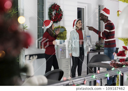 Smiling diverse business team socialising by office water cooler, discussing Christmas holiday season plans. Relaxed coworkers enjoying nice chat during coffee break in festive decorated workspace 109187462