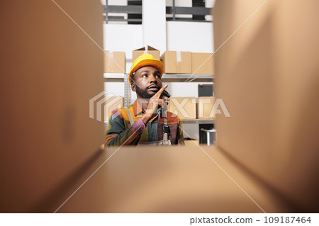 Selective focus of employee checking cardboard boxes, working at clients orders preparing packages for delivery in warehouse. African american stockroom worker wearing industrial overall and helmet 109187464