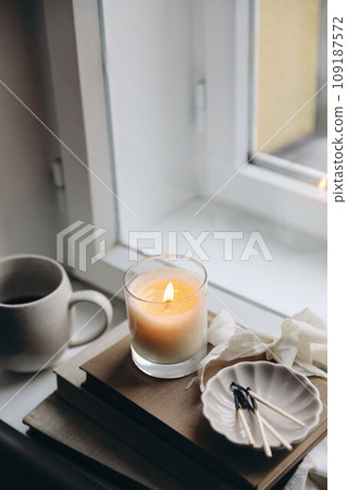 Cozy autumn, winter still life. Burning candle on old books. Matches, ceramic plate on windowsill. Blurred cup of coffee, mug. Moody fall setting. Thanksgiving. Halloween. Blurred vertical top view. 109187572