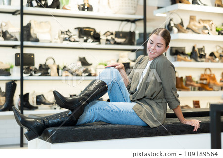 Young woman trying on pair of black cossack boots at footwear boutique 109189564