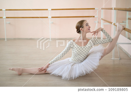 Beautiful ballerina in the body and white tutu practicing in dance class and sitting in the splits. Young flexible dancer posing in pointe shoes. 109189805