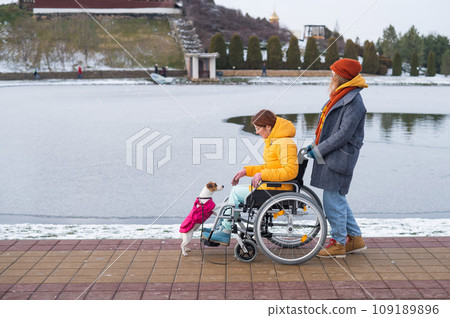 Caucasian woman driving her friend in a wheelchair along the lake in winter. Caucasian woman driving her friend in a wheelchair along the lake in winter. 109189896