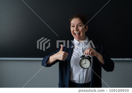 Happy female teacher in pantsuit holding an alarm clock standing at the school board and showing thumbs up. Happy female teacher in pantsuit holding an alarm clock standing at the school board and showing thumbs up. 109189930