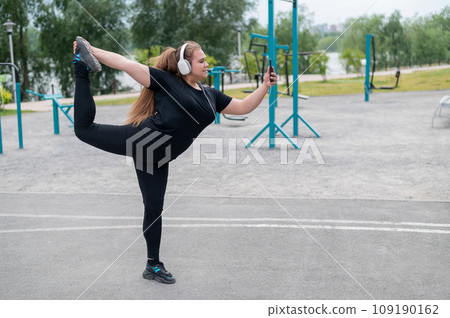 Beautiful fat young woman in headphones makes a figure skater and takes a selfie. A chubby smiling girl listens to music and warms up before jogging outdoors. Sports for weight loss. Beautiful fat young woman in headphones makes a figure skater and takes a selfie. A chubby smiling girl listens to music and warms up before jogging outdoors. Sports for weight loss. 109190162