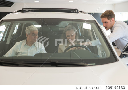A gray-haired Caucasian woman is sitting behind the wheel of a car. Car dealership salesman consulting mature married couple. A gray-haired Caucasian woman is sitting behind the wheel of a car. Car dealership salesman consulting mature married couple. 109190180
