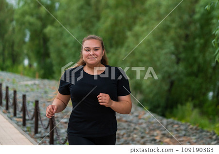 An obese young woman jogging outdoors. Fat beautiful smiling girl in a black tracksuit is engaged in fitness for weight loss on the waterfront. A woman runs on a summer day. An obese young woman jogging outdoors. Fat beautiful smiling girl in a black tracksuit is engaged in fitness for weight loss on the waterfront. A woman runs on a summer day. 109190383