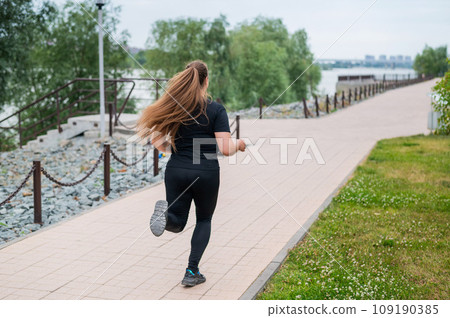 An obese young woman jogging outdoors. Fat beautiful smiling girl in a black tracksuit is engaged in fitness for weight loss on the waterfront. A woman runs on a summer day. An obese young woman jogging outdoors. Fat beautiful smiling girl in a black tracksuit is engaged in fitness for weight loss on the waterfront. A woman runs on a summer day. 109190385