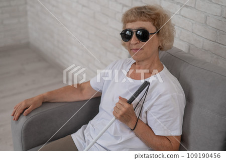An elderly blind woman wearing sunglasses and with a tactile cane sits on the sofa. An elderly blind woman wearing sunglasses and with a tactile cane sits on the sofa. 109190456