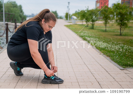 A fat woman in a tracksuit crouches down and ties her shoelaces outdoors. 109190799
