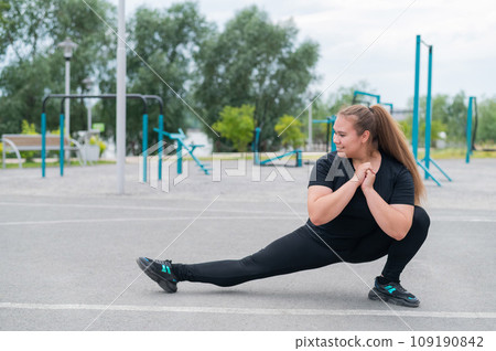 Obese young caucasian woman doing side lunges. Fat beautiful smiling girl in a black tracksuit is engaged in fitness outdoors. Warming up before training. 109190842