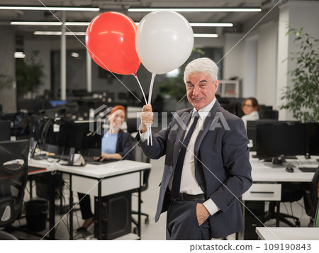 Portrait of a cheerful mature business man holding balloons in the office. 109190843
