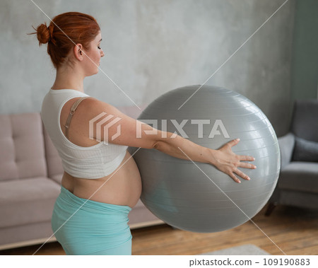 Pregnant red-haired woman doing arm exercises with fitball at home. Pregnant red-haired woman doing arm exercises with fitball at home. 109190883