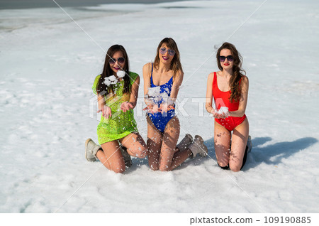 Three girlfriends in swimsuits are relaxing on a snow-covered beach. Hot girls posing in bikinis outdoors in winter.  109190885