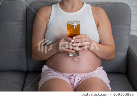 Close-up of the belly of a pregnant woman holding a glass of beer while sitting on the sofa. 109190886