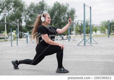 A beautiful fat girl in headphones is engaged in fitness on the sports ground and takes a selfie. Young woman lunges and is photographed on a smartphone outdoors on a warm summer day. 109190922