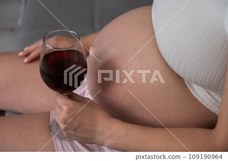 Close-up of the belly of a pregnant woman holding a glass of red wine while sitting on the sofa.  109190964