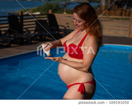 A red-haired pregnant woman sunbathes by the pool. The expectant mother applies sunscreen to her tummy. 109191050