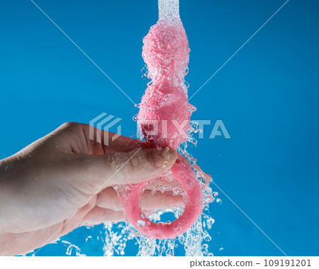 Woman holding pink anal beads under running water on blue background. Sex toy hygiene concept. Woman holding pink anal beads under running water on blue background. Sex toy hygiene concept. 109191201