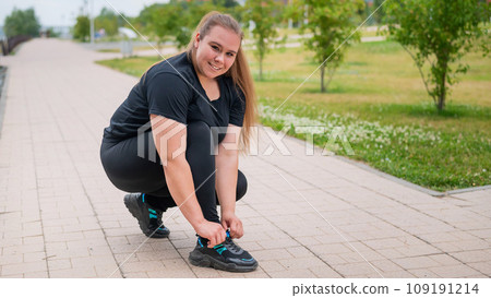 A fat woman in a tracksuit crouches down and ties her shoelaces outdoors. A fat woman in a tracksuit crouches down and ties her shoelaces outdoors. 109191214