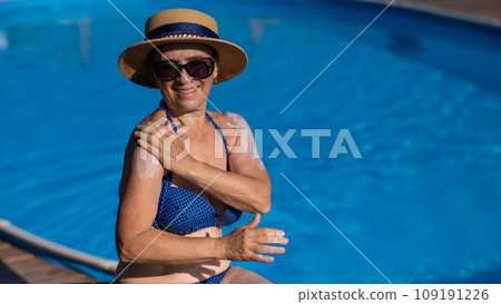 Portrait of an old woman in a straw hat, sunglasses and a swimsuit applying sunscreen to her skin while relaxing by the pool.  109191226