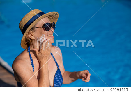Portrait of an old woman in a straw hat, sunglasses and a swimsuit applying sunscreen to her face while relaxing by the pool.  109191315