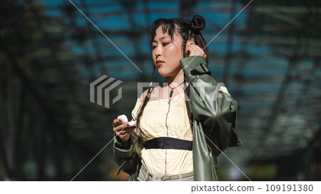 A beautiful Asian woman in shorts and a green leather coat comes out of the subway and uses wireless headphones. A beautiful Asian woman in shorts and a green leather coat comes out of the subway and uses wireless headphones. 109191380