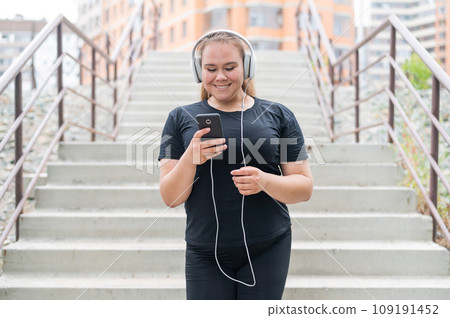 A fat young woman goes down the stairs and listens to music on a smartphone. Obese girl with headphones resting after jogging. 109191452