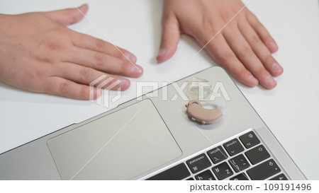 Close-up of a man's hands with a hearing aid near a laptop. Close-up of a man's hands with a hearing aid near a laptop. 109191496
