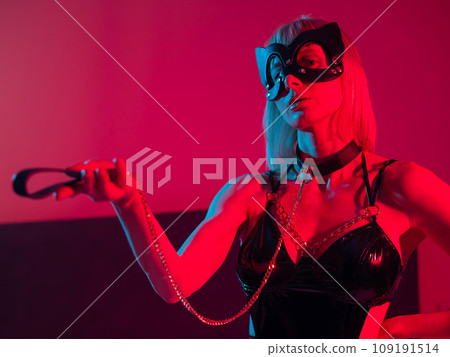 Blonde girl in a leather mask and handcuffs in the bedroom in red blue neon light. Role-playing games. Blonde girl in a leather mask and handcuffs in the bedroom in red blue neon light. Role-playing games. 109191514