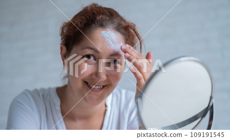 Portrait of a Caucasian woman with vitiligo uses sunscreen. A girl with a white pigment spot on her forehead looks in the mirror and is smeared with cream. 109191545