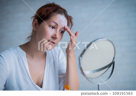 Portrait of a Caucasian woman with vitiligo uses sunscreen. A girl with a white pigment spot on her forehead looks in the mirror and is smeared with cream. 109191563