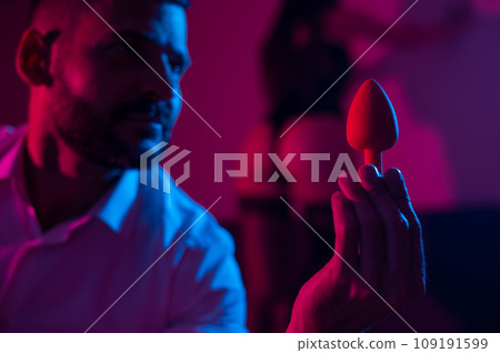 Bearded caucasian man holding an anal plug. Close-up of female buttocks in underwear. Sexual preferences of the couple. 109191599