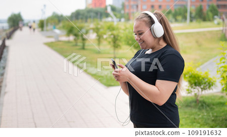 A beautiful fat girl in black sportswear listens to music on headphones and text messages on the phone while walking outdoors. A plump young woman is leafing through a music playlist in smartphone. 109191632