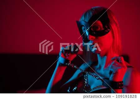 Blonde girl in a leather mask and handcuffs in the bedroom in red blue neon light. Role-playing games. Blonde girl in a leather mask and handcuffs in the bedroom in red blue neon light. Role-playing games. 109191828