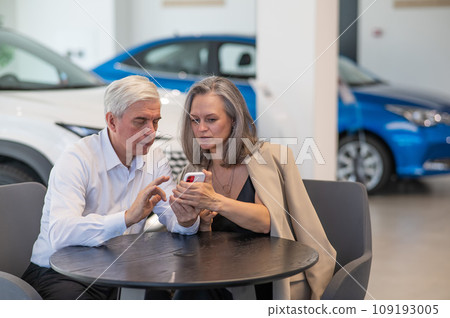 A mature couple is sitting in a car dealership and looking at a smartphone. A mature couple is sitting in a car dealership and looking at a smartphone. 109193005