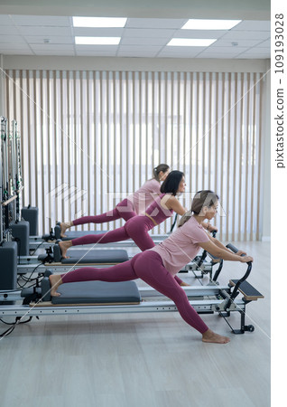 Three Asian women in pink sportswear do exercises on the reformer machine. Pilates classes. Three Asian women in pink sportswear do exercises on the reformer machine. Pilates classes. 109193028