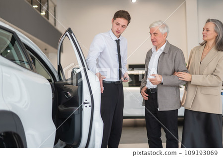 A salesman in a car dealership shows a car to a mature married couple. 109193069