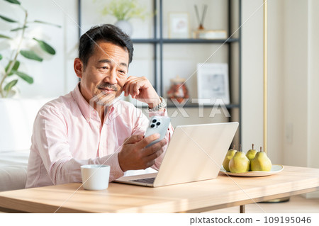 一位老人在客廳使用筆記型電腦和智慧型手機。 109195046