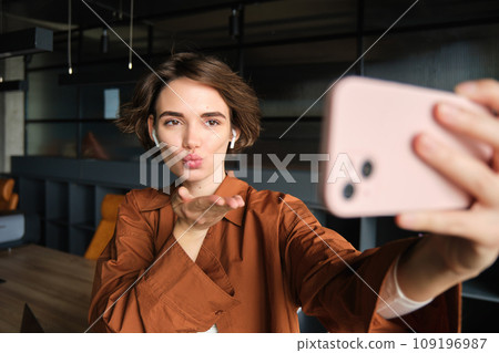 Portrait of woman taking selfie in office, wearing casual clothes, posing in wireless headphones, making photos on mobile phone Portrait of woman taking selfie in office, wearing casual clothes, posing in wireless headphones, making photos on mobile phone 109196987