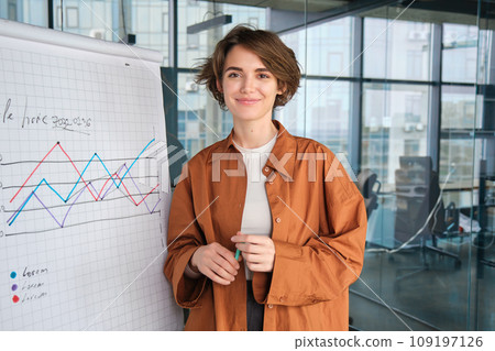 Image of young saleswoman, start up manager stands near board with diagram, giving presentation to team in office and smiling 109197126