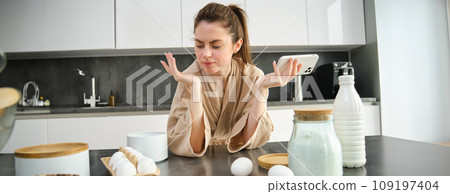 Attractive young cheerful girl baking at the kitchen, making dough, holding recipe book, having ideas Attractive young cheerful girl baking at the kitchen, making dough, holding recipe book, having ideas 109197404